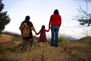 A still from the New Day film Sin Pais. The backs of three family members holding hands, a small child in the middle. They walk on a sandy path with tall grass on either side.
